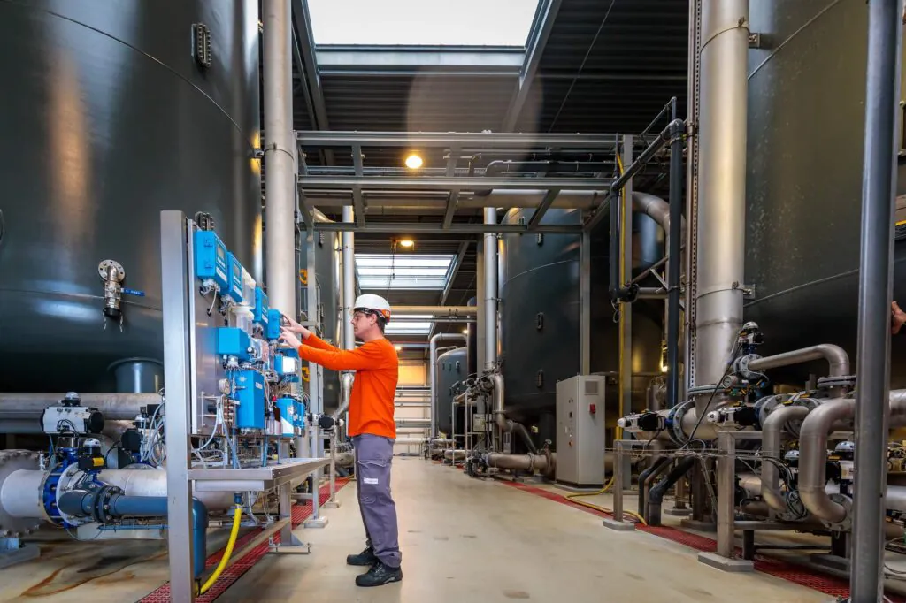 Een technicus in werkkleding bedient een bedieningspaneel in een industriële hal met grote opslagtanks en complexe pijpleidingen, wat bijdraagt aan effectief asset management in deze technische omgeving.