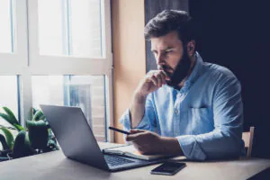 Een man met een baard zit geconcentreerd achter zijn laptop aan een bureau bij een raam, terwijl hij werkt aan effectief asset management in een kantooromgeving.