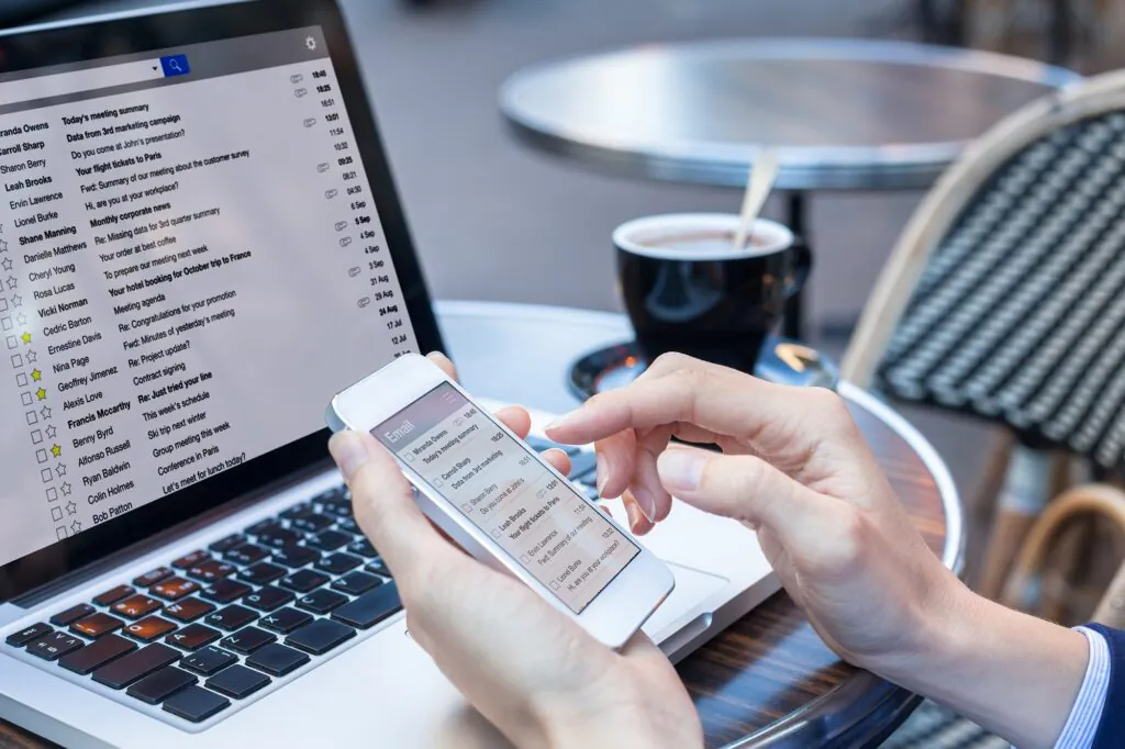 Iemand gebruikt een smartphone om e-mails te beheren, wat essentieel is voor efficiënt **field service management**, terwijl een laptop en koffie op een terras staan. **Tekst op het scherm:** Miranda Owens Today's meeting summary 18:45 Carroll Sharp Data from 3rd marketing 16:51 Sharon Berry Do you come at John's 13:01 Leah Brooks Your flight tickets to Paris 11:54 Ervin Lawrence Fwd: Summary of our meeting 11:32 Lionel Burke Hi, are you at your workplace? 10:32 (Laptop scherm bevat vergelijkbare e-maillijsten en namen als: Miranda Owens, Carroll Sharp, Sharon Berry, Leah Brooks, Ervin Lawrence, Lionel Burke, Shane Manning, Danielle Matthews, Cheryl Young, Rosa Lucas, Vicki Norman, Cedric Barton, Ernestine Davis, Nina Page, Geoffrey Jimenez, Alexis Love, Francis McCarthy, Benny Byrd, Alfonso Russell, Ryan Baldwin, Colin Holmes, Bob Patton).