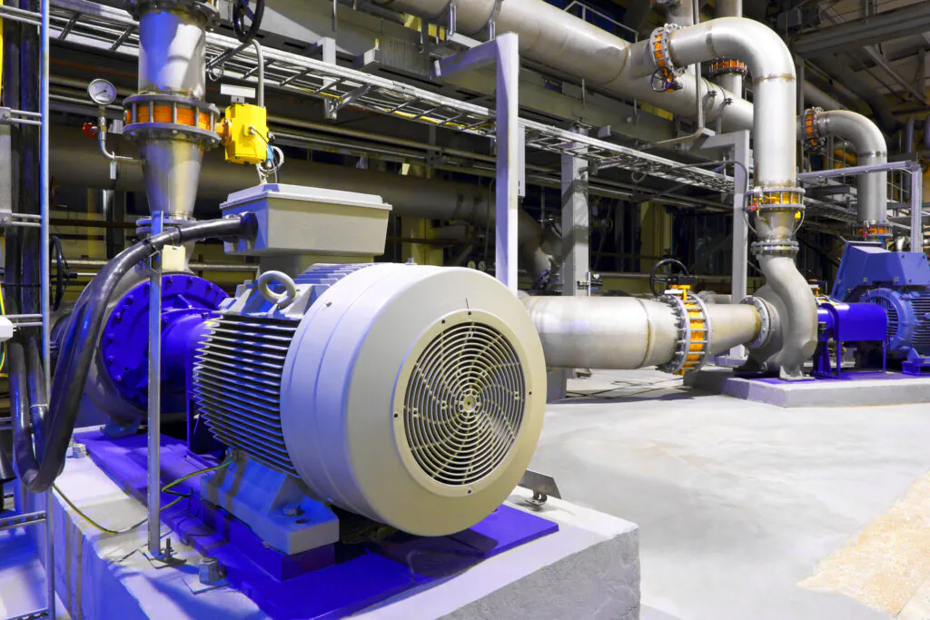 Een industriële elektromotor en bijbehorende pomp staan opgesteld in een fabriekshal, een essentieel onderdeel voor efficiënt asset management in een technische omgeving met complexe leidingwerk- en installatiestructuren.