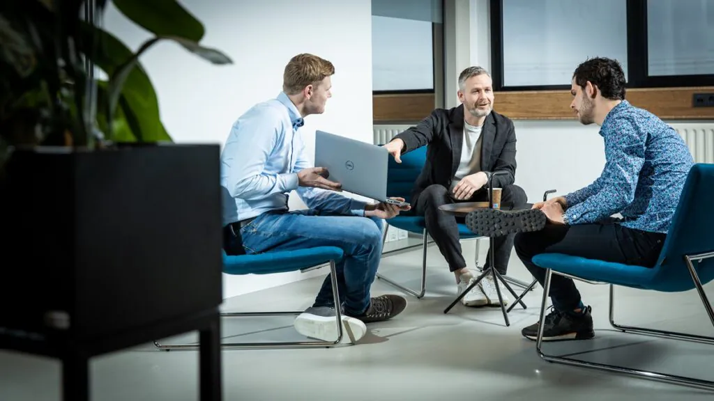 Drie mannen bespreken werkgerelateerde zaken op een laptop in een kantooromgeving; ze bespreken mogelijk SAP FSM-oplossingen voor efficiënt beheer terwijl ze op blauwe stoelen in een lichte ruimte zitten.