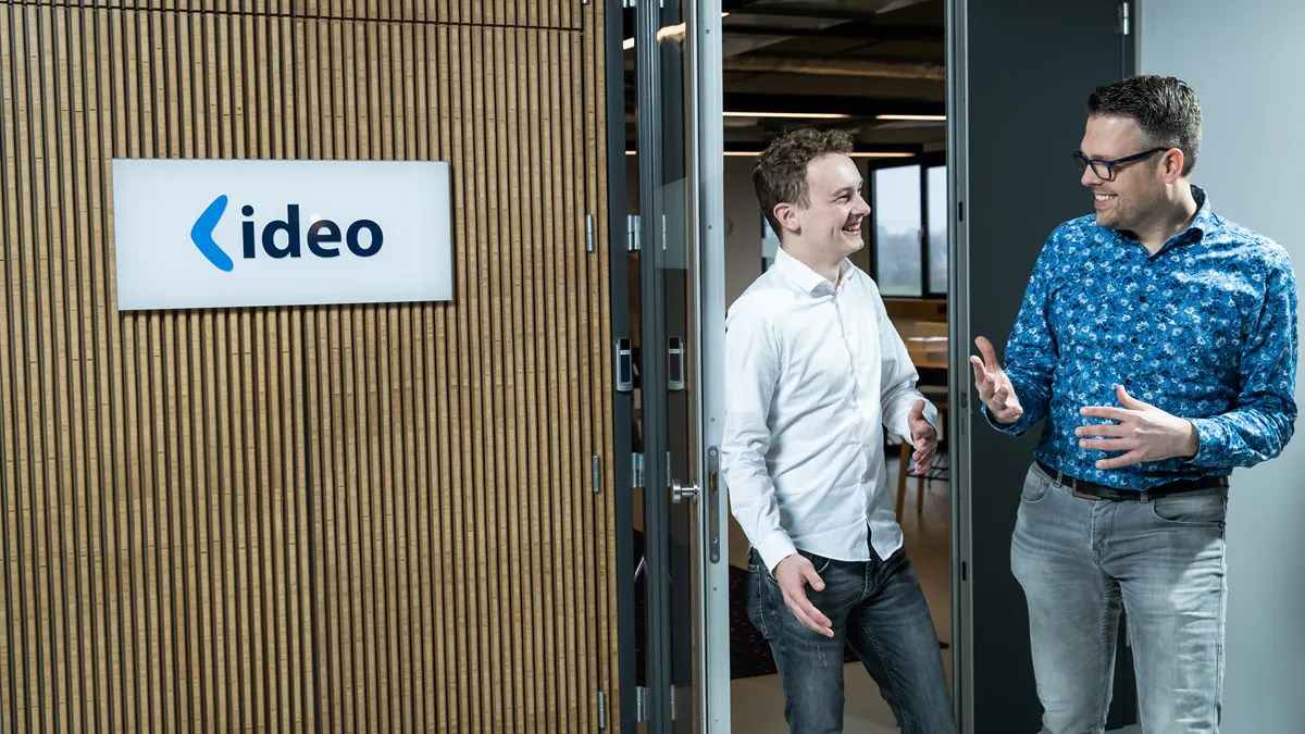 Twee mannen praten en glimlachen bij een kantooringang met een "ideo" logo aan de wand, wat symbool staat voor de professionele samenwerking binnen oplossingen voor sap fsm.