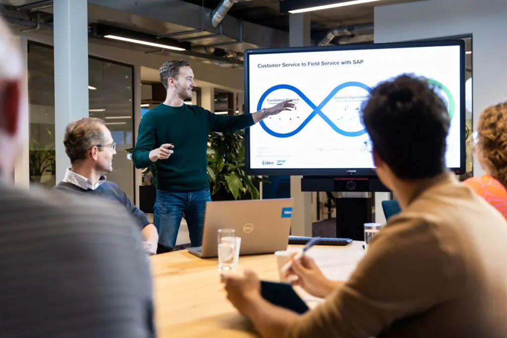 Een spreker presenteert een procesflow over field service management op een groot scherm voor een groep collega's in een modern kantoor. Tekst op scherm: "Customer Service to Field Service with SAP", "Customer Service", "Service Operations", "ideo SAP", "CTOUCH".
