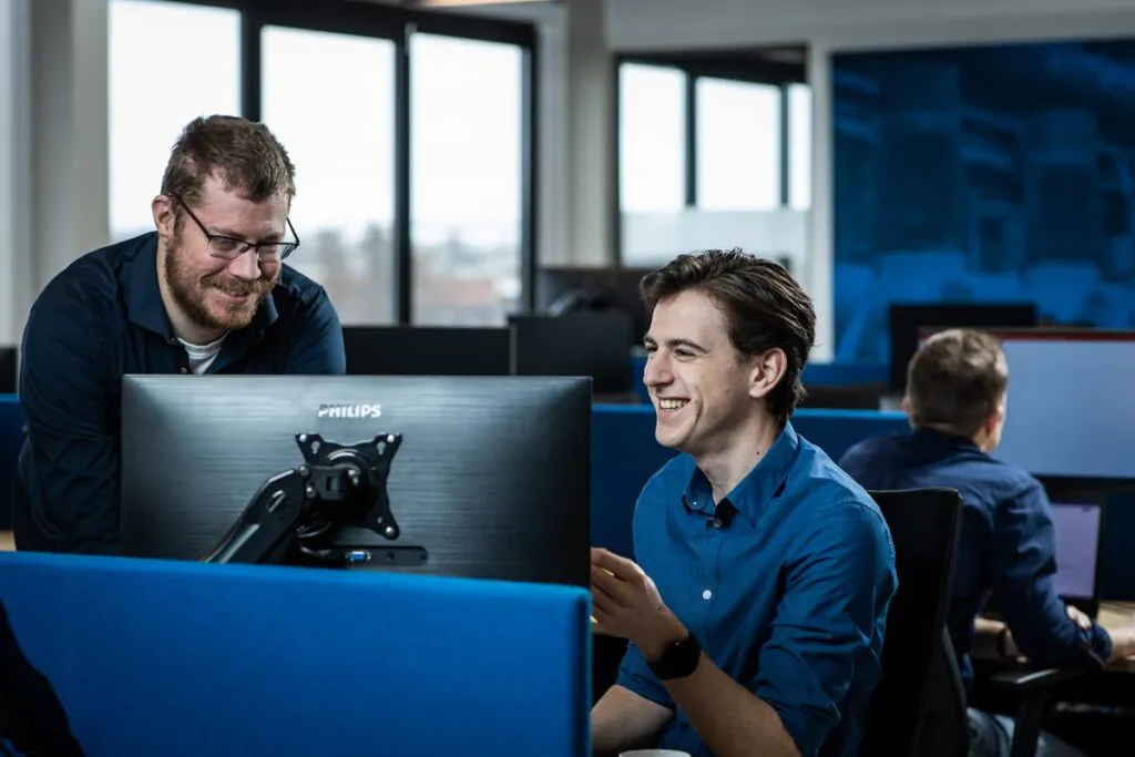 Twee collega's lachen naar een Philips-monitor in een modern kantoor, waarbij ze samenwerken aan softwareoplossingen gerelateerd aan field service management om de bedrijfsprocessen te optimaliseren.