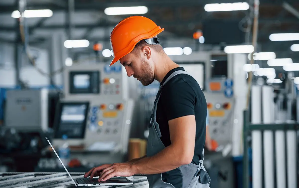 Een technicus met een oranje helm werkt op een laptop voor effectief asset management in een drukke fabriekshal met industriële machines op de achtergrond.