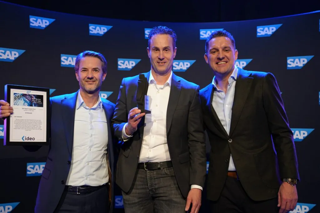 Drie mannen in pak lachen terwijl ze een trofee en oorkonde vasthouden op een SAP-evenement, waarbij ze hun expertise in asset management en SAP technology vieren voor een merkachtergrond. Tekst op de oorkonde: "SAP Partner Awards 2022 GAMECHANGERS SAP Digital Supply Chain Partner of the Year WINNAAR JURY RAPPORT Wanneer het gaat over Asset Management als ook Field Service Management, dan kun je niet om Ideo heen. Dankzij hun uitstekende expertise en jarenlange ervaring op dit gebied, de organisatie van meerdere webinars voor business users en met als kern op de kaart het Service Management in de praktijk event in het Ideo Experience Center - waar zowel Eneco als Van Oestel hun klantervaringen deelden - kozen Brabant Water, Vitens, SPIE als ook Boon Edam voor SAP Digital Supply Chain." "<Ideo