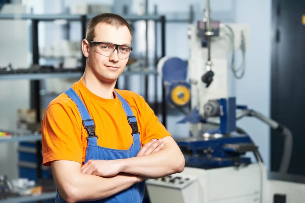 Een technicus in werkkleding met een veiligheidsbril poseert zelfverzekerd met zijn armen over elkaar in een werkplaats, ondersteund door geavanceerde SAP FSM-oplossingen voor efficiënt field service management.