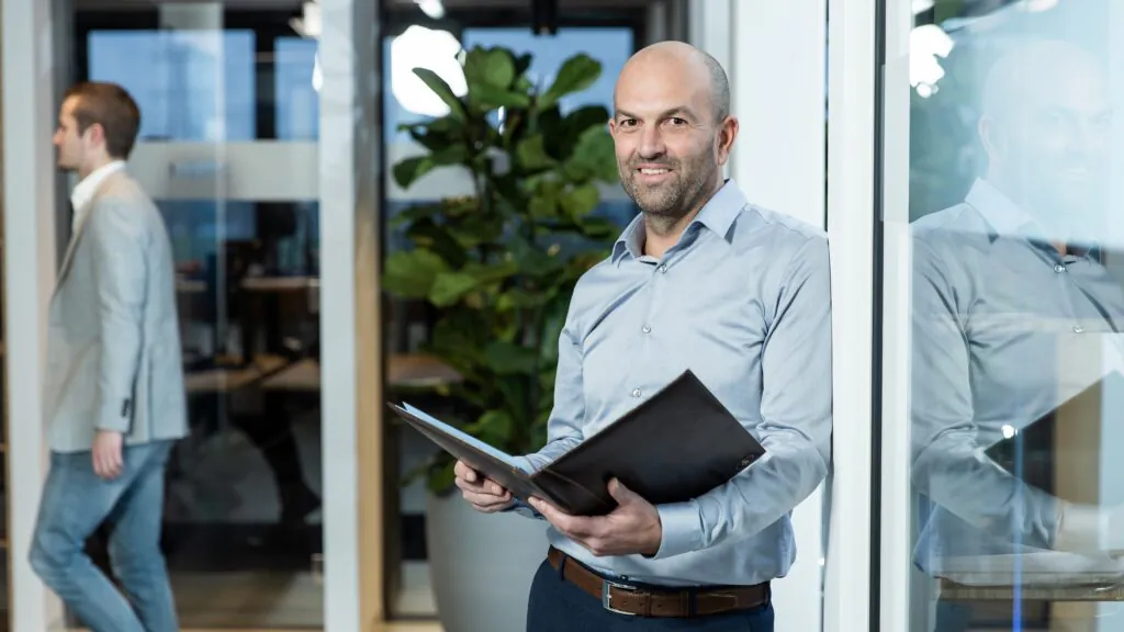 Een glimlachende man in een blauw hemd houdt een map vast terwijl hij leunt tegen een glazen wand in een kantooromgeving, geoptimaliseerd voor efficiënt asset management.