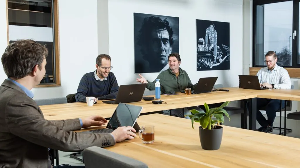 Vier professionele mannen werken op laptops aan een grote houten tafel in een modern kantoor, waarbij ze strategieën voor asset management bespreken terwijl er koffie en thee klaarstaat.