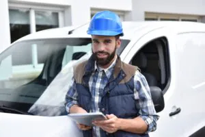 Een vakman met een blauwe helm en tablet staat voor zijn witte bestelbus en glimlacht, wat essentieel is voor efficiënt field service management bij technische werkzaamheden op locatie.