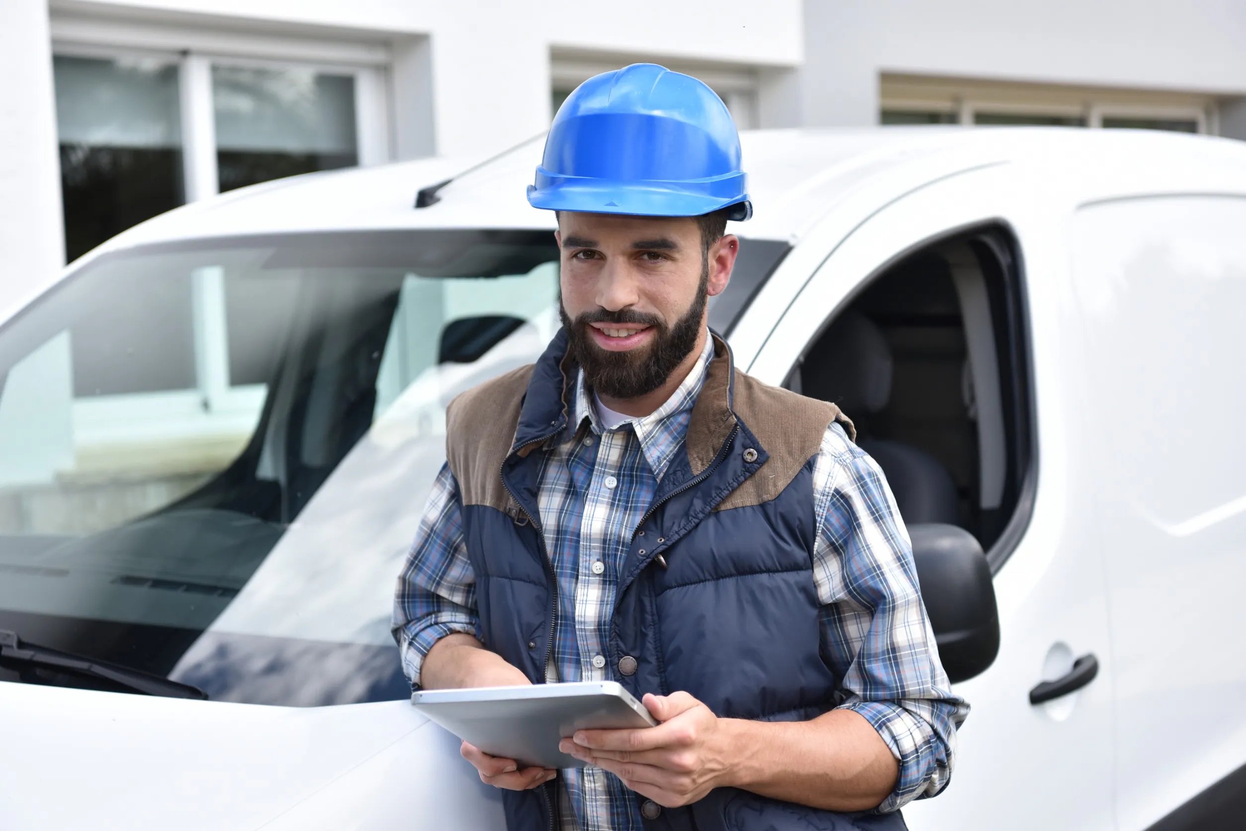 Een vakman met een blauwe helm en tablet staat voor zijn witte bestelbus en glimlacht, wat essentieel is voor efficiënt field service management bij technische werkzaamheden op locatie.