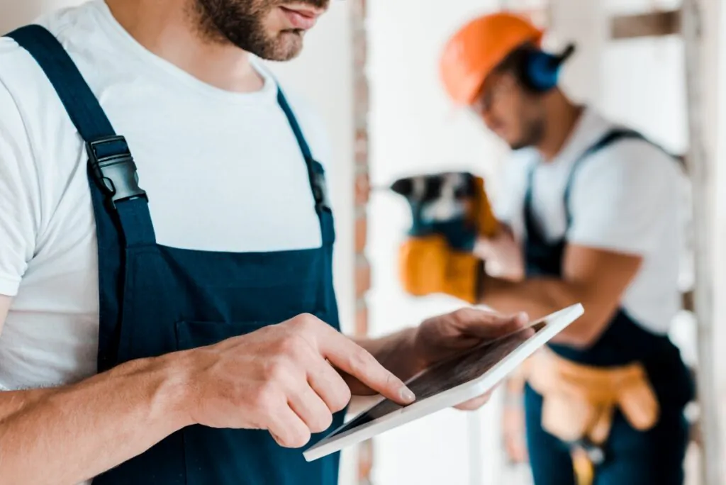 Een bouwvakker in werkkleding bedient een tablet voor SAP FSM, terwijl op de achtergrond een collega met een boormachine in een bouwomgeving werkt.