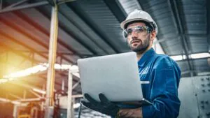 Een technicus in werkkleding, helm en veiligheidsbril bekijkt een laptop voor efficiënt field service management in een grote, verlichte fabriekshal met stalen constructies op de achtergrond.