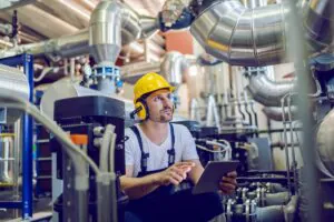 Een technicus met helm en gehoorbescherming gebruikt een tablet om apparatuur te inspecteren voor efficiënt field service management in een industriële omgeving vol met metalen leidingen en machines.