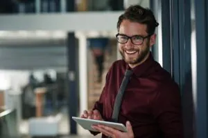 Een lachende man met een bril en bordeauxrood overhemd bedient een tablet in een modern kantoorpand, ideaal voor het beheren van processen zoals field service management.
