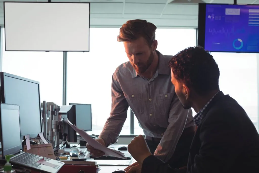 Twee professionals bespreken documenten op een modern kantoor, waarbij ze samenwerken aan strategieën voor field service management met digitale dashboards en schermen op de achtergrond.