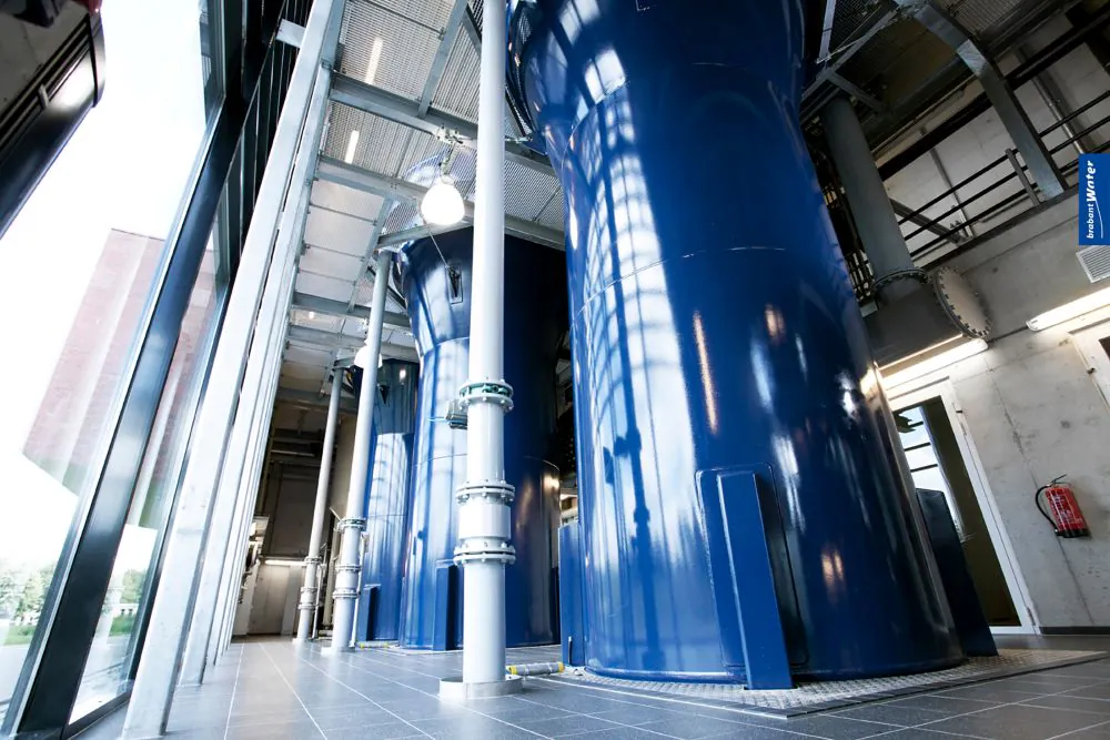 Grote, glanzende blauwe industriële tanks staan in een moderne hal en ondersteunen efficiënt asset management, geflankeerd door een glazen wand en betonnen constructies. Tekst in de afbeelding: brabant Water.