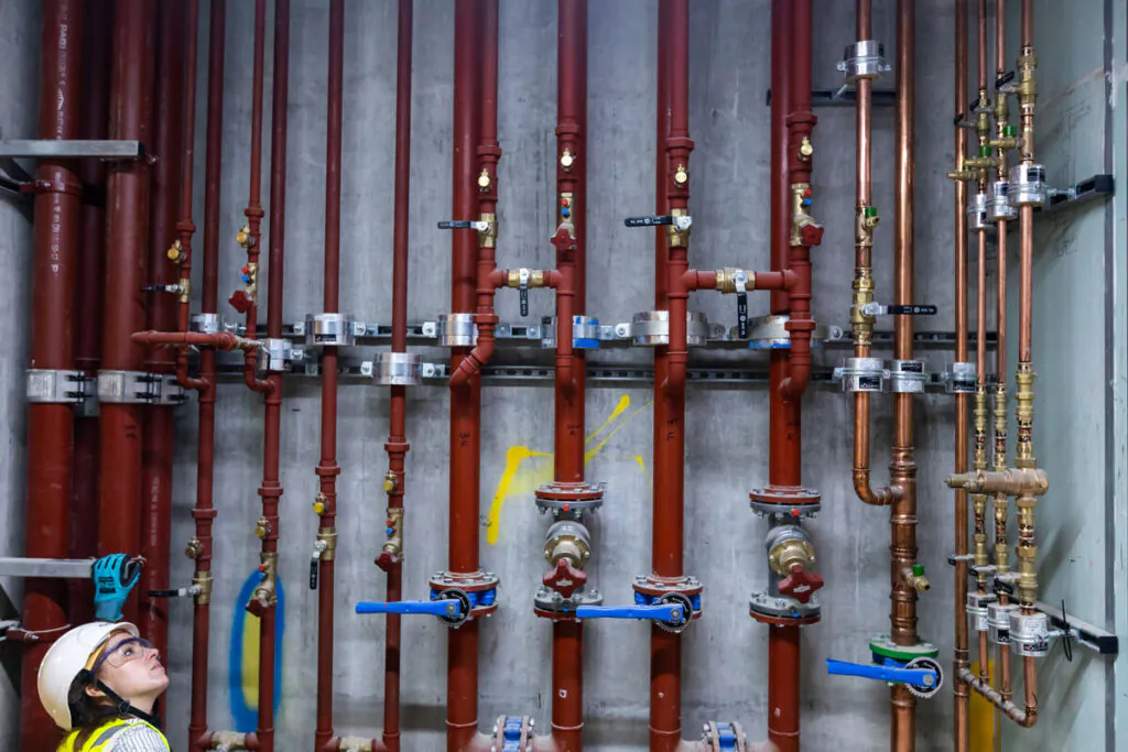 Een technicus inspecteert met field service management een netwerk van verticale rode en koperen leidingen met diverse kranen en koppelingen, gemonteerd op een industriële betonnen muur.