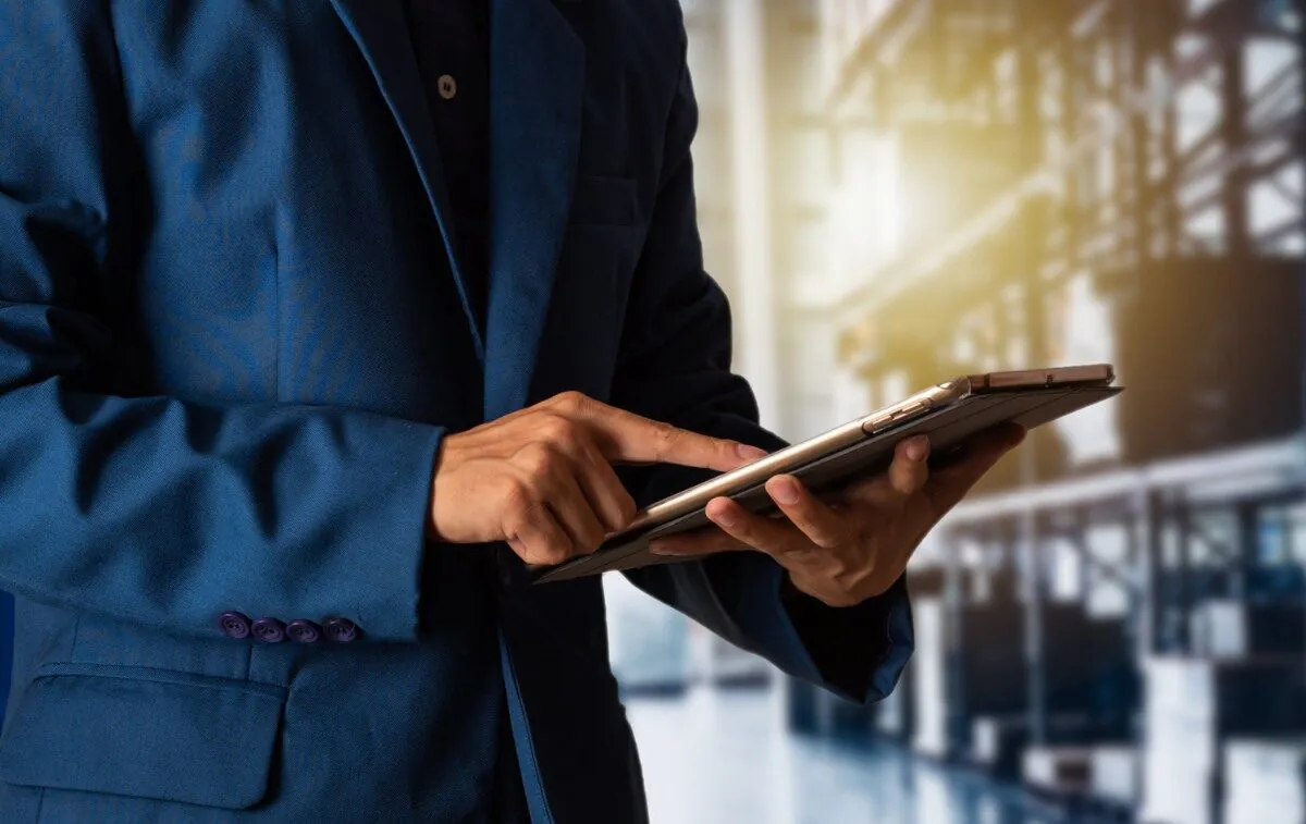 Een professional in een blauw jasje bedient een tablet voor efficiënt asset management, terwijl hij zich in een wazig magazijn vol opslagrekken bevindt.