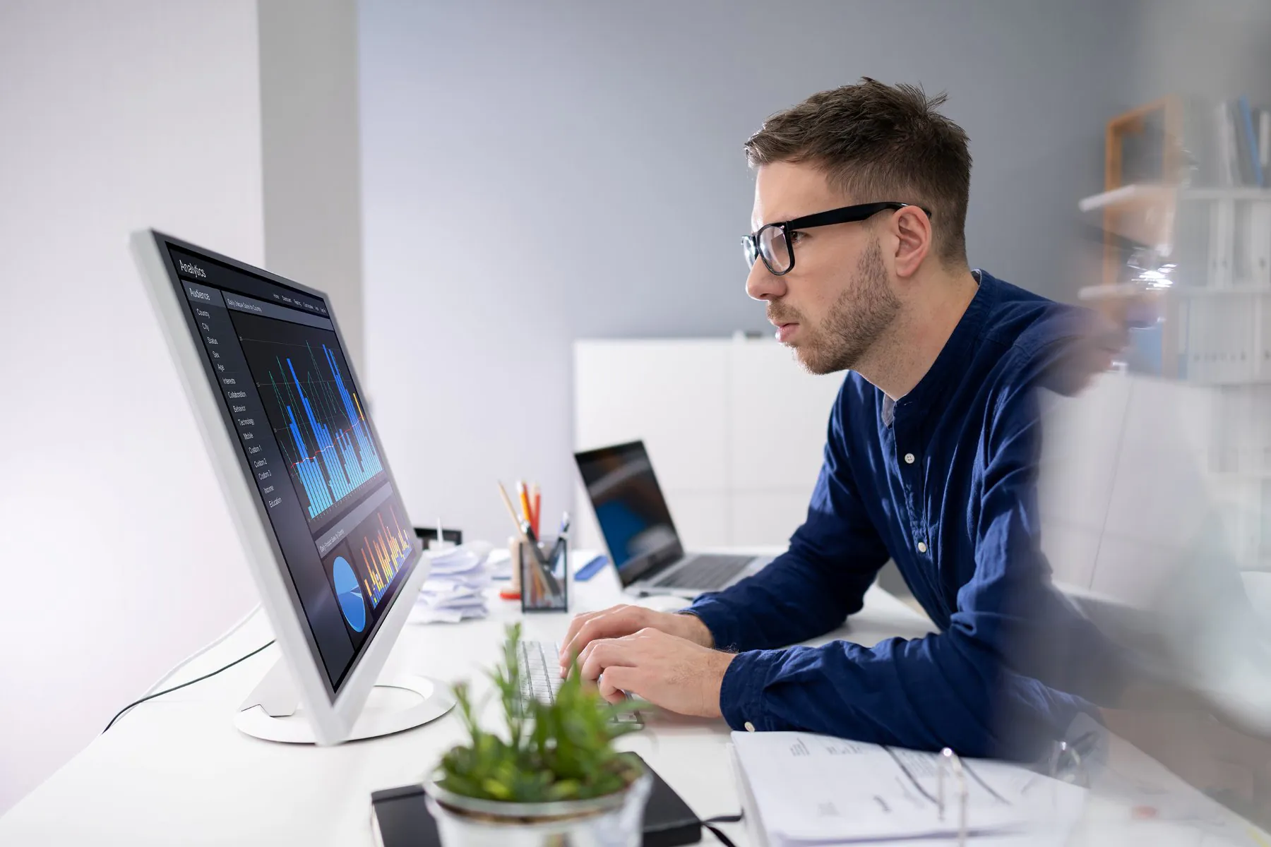 Een man met een bril typt op een computer in een kantoor, waarbij hij data over asset management analyseert op een scherm met grafieken en tabellen.