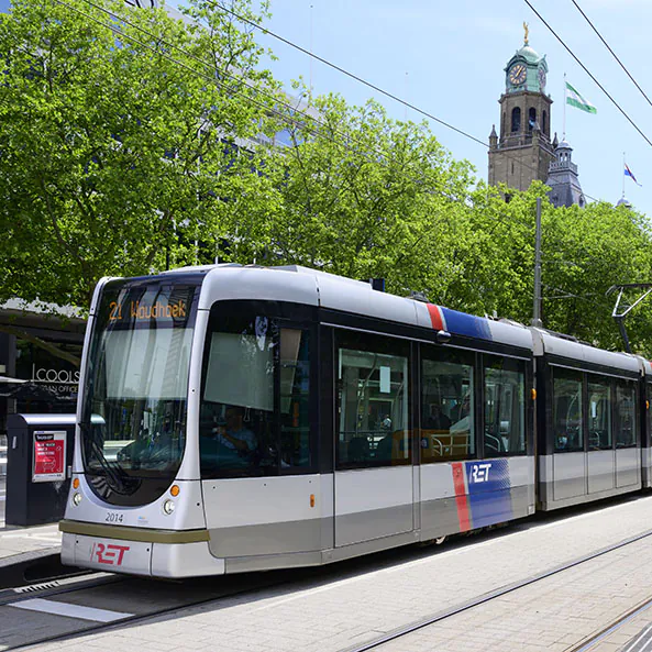 Een moderne RET-tram rijdt over rails door een stedelijke straat in Rotterdam, ondersteund door effectief asset management, met op de achtergrond bomen en de historische toren van het stadhuis. Tekst op de tram: "21 Woudhoek", "2014", "RET".