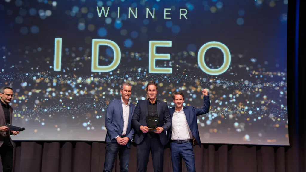 Drie mannen in pak vieren op een podium hun prijs voor field service management voor een scherm met de tekst: WINNER IDEO.