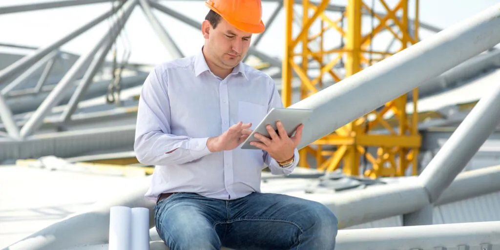 Een bouwopzichter in een oranje helm bedient een tablet voor efficiënt field service management terwijl hij op een stalen constructie van een bouwplaats zit.