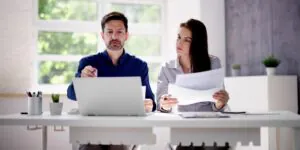 Twee professionals werken samen aan een laptop en documenten op een licht kantoor, waarbij ze zich focussen op efficiënt asset management ter ondersteuning van hun professionele werkzaamheden.