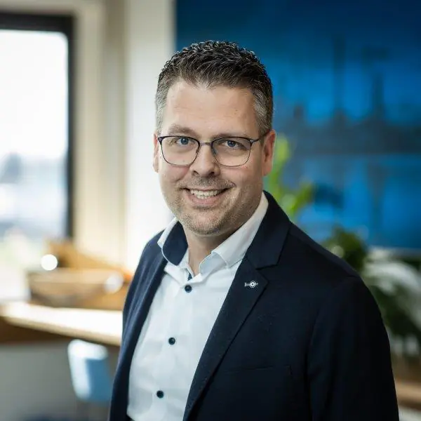 Een professionele man met bril glimlacht in een kantooromgeving en is een expert in field service management, terwijl hij op de achtergrond een wazig blauw interieur heeft.